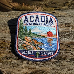 May include: A rectangular embroidered patch featuring a landscape of Acadia National Park. The design shows green pine trees, rocky cliffs, a blue ocean, and a red sun. Text reads ACADIA NATIONAL PARK and MAINE EST. 1919 with a small green tree icon.