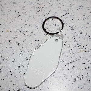 May include: A white, retro-style motel keychain with a silver edge and a black keyring. The keychain has a smooth, blank surface, and the background is a speckled white and black pattern.