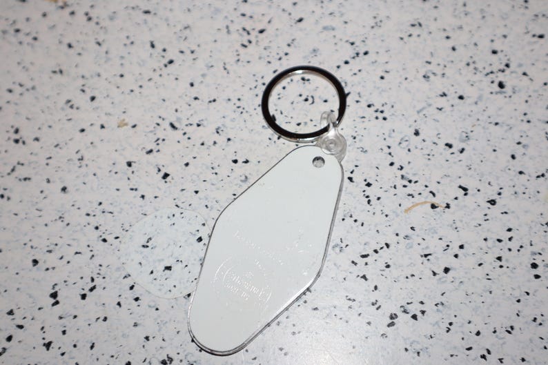 May include: A white, retro-style motel keychain with a silver edge and a black keyring. The keychain has a smooth, blank surface, and the background is a speckled white and black pattern.