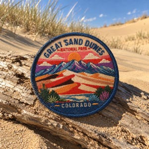 May include: A circular embroidered patch featuring a landscape of Great Sand Dunes National Park in Colorado. The design includes a bright orange sun, purple mountains, tan sand dunes, and green desert plants, with the text Great Sand Dunes National Park and Colorado.