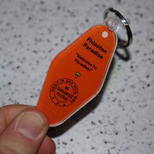 Op de afbeelding: Oranje plastic motel sleutelhanger met de tekst "Fhloston Paradise" en "Welcome To Paradise!"  Aan de achterkant van de sleutelhanger staat een zwart-wit stempel met de tekst "Drop in any mail box. We guarantee postage."