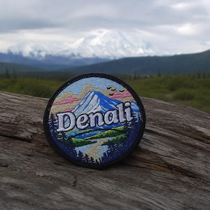 May include: A circular embroidered patch featuring a mountain landscape with a snow-capped peak, green trees, and a river. The word Denali is stitched in white capital letters across the center of the design, framed by a black border.