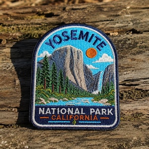 May include: An embroidered patch featuring a landscape of Yosemite National Park. The design includes a large granite mountain, a waterfall, green pine trees, a blue river, and an orange sun in a blue sky. Text reads YOSEMITE NATIONAL PARK CALIFORNIA.