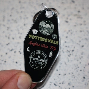 Op de afbeelding: Zwart-witte motel sleutelhanger met de tekst "POTTERSVILLE Bedford Falls, NY" en een kleine illustratie van een stad met een halve maan. De tekst "DROP IN ANY MAIL BOX WE GUARANTEE POSTAGE" is rond een ster gedrukt.