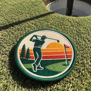 May include: A circular embroidered patch featuring a golfer in a dark green silhouette swinging a club. The background displays a sunset with horizontal stripes of yellow, orange, and red, two dark green pine trees, and a red golf flag.