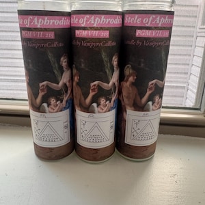 May include: Three glass jar candles with a dark brown wax. Each candle has a label with the text "Stele of Aphrodite" and "PGM VII. 215 by VampyreCallisto". The label features a classical art scene with figures and text.