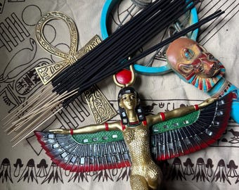 May include: A bundle of black incense sticks resting on a gold-colored ankh, a blue circular object, a small lion-headed figurine, and a kneeling winged goddess statue with a red sun disk on her head, featuring gold, green, and black details.