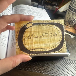 Puede incluir: Un trozo rectangular de papiro con un diseño de ouroboros negro que rodea una escritura antigua. El papiro se sostiene contra una superficie oscura, con un libro y otros objetos en el fondo.