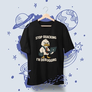 May include: Black t-shirt featuring a cartoon duck wearing a tie and holding a laptop and briefcase. The text on the shirt reads "STOP QUACKING I'M DEBUGGING". The design is set against a light blue background with space-themed doodles.
