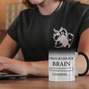 Op de afbeelding: Een zwart-witte mok die van kleur verandert met de tekst "PROGRAMMER BRAIN LOADING...". De mok is zwart als hij koud is en wordt wit als hij warm is. Op de achtergrond gebruikt een persoon een laptop.