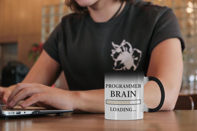 Op de afbeelding: Een zwart-witte mok die van kleur verandert met de tekst "PROGRAMMER BRAIN LOADING...". De mok is zwart als hij koud is en wordt wit als hij warm is. Op de achtergrond gebruikt een persoon een laptop.