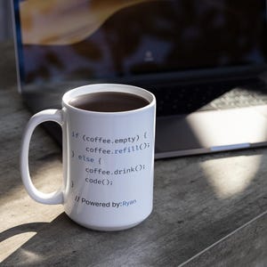 May include: White coffee mug filled with dark liquid, likely coffee, with a handle. The mug has code printed on it, including "if (coffee.empty)" and "Powered by: Ryan". A laptop is in the background.