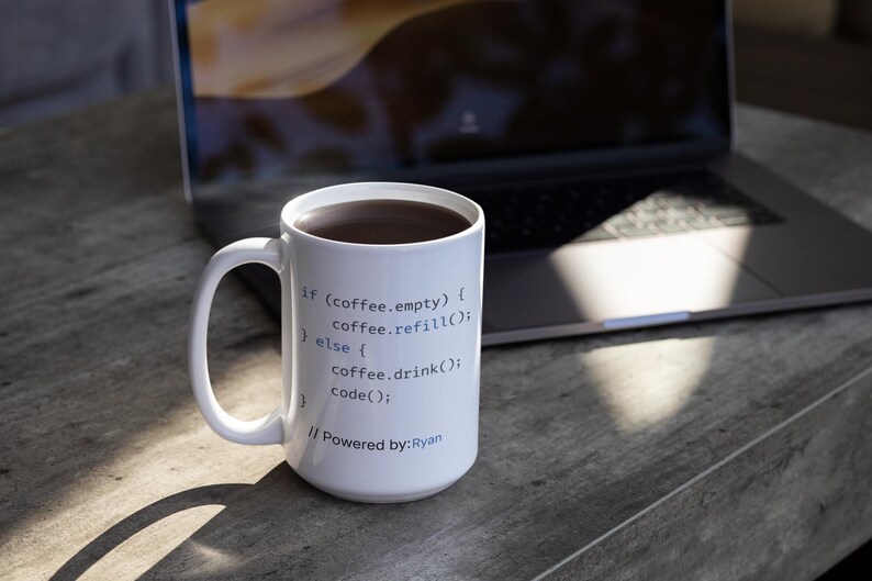 May include: White coffee mug filled with dark liquid, likely coffee, with a handle. The mug has code printed on it, including "if (coffee.empty)" and "Powered by: Ryan". A laptop is in the background.
