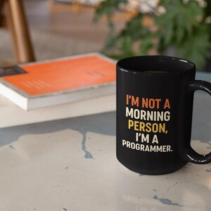 Könnte beinhalten: Schwarze Kaffeetasse mit dem Text "I'M NOT A MORNING PERSON, I'M A PROGRAMMER." Die Tasse steht auf einem hellblauen Tisch neben zwei Büchern. Das obere Buch hat einen orangefarbenen Einband.
