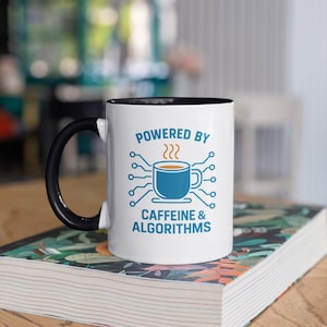 May include: White coffee mug with a black handle and interior, featuring the text "POWERED BY CAFFEINE & ALGORITHMS" in blue. The mug has a blue coffee cup graphic with steam rising above it. The mug is sitting on a book.