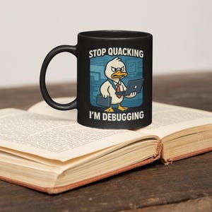Peut inclure: Mug noir avec un canard de dessin animé portant une cravate et tenant un ordinateur portable. Le mug affiche le texte "STOP QUACKING I'M DEBUGGING" en blanc. Le mug est posé sur un livre ouvert.