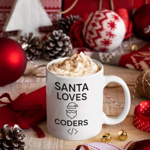 Peut inclure: Une tasse en céramique blanche avec le texte "SANTA LOVES CODERS" et un dessin du Père Noël. La tasse est remplie d'une boisson garnie de crème fouettée. Des décorations de Noël, dont des boules et des pommes de pin, entourent la tasse.