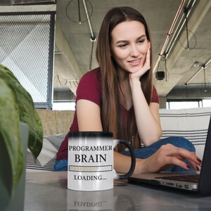 Op de afbeelding: Een zwart-wit veranderende mok met de tekst "PROGRAMMER BRAIN LOADING..." staat op een tafel. Een persoon zit voor een laptop, met hun hand op hun kin. De persoon draagt een bordeauxrode shirt en een spijkerbroek.