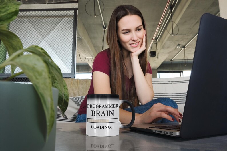 Op de afbeelding: Een zwart-wit veranderende mok met de tekst "PROGRAMMER BRAIN LOADING..." staat op een tafel. Een persoon zit voor een laptop, met hun hand op hun kin. De persoon draagt een bordeauxrode shirt en een spijkerbroek.
