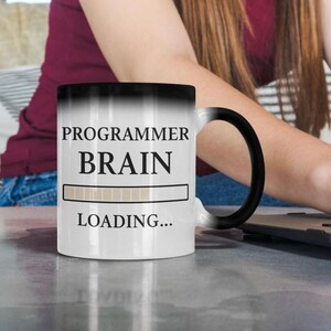 Op de afbeelding: Witte keramische mok met een zwart handvat, met de tekst "PROGRAMMER BRAIN LOADING..." in zwart. De mok heeft een laadbalkgrafiek. De mok staat op een grijs oppervlak.