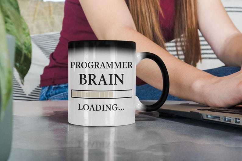 Op de afbeelding: Witte keramische mok met een zwart handvat, met de tekst "PROGRAMMER BRAIN LOADING..." in zwart. De mok heeft een laadbalkgrafiek. De mok staat op een grijs oppervlak.