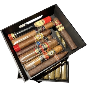 May include: A black wooden box filled with various cigars, including The Traveler, Rock House, and a few others. The box is stacked on top of another box filled with more cigars.