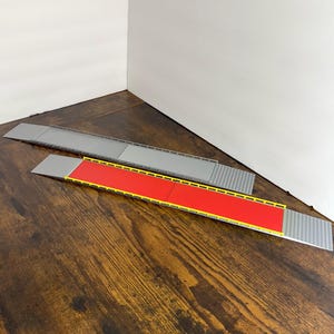 May include: Two plastic toy ramps. One is gray, the other is red with yellow trim. Both have a corrugated surface on the ends and are designed to connect to each other. The ramps are on a wooden surface.