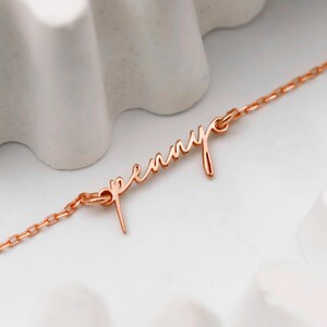 May include: A delicate rose gold necklace with the name "Penny" in cursive script. The chain is fine and links to the name pendant. The necklace is displayed on a white surface with a textured, wave-like background.