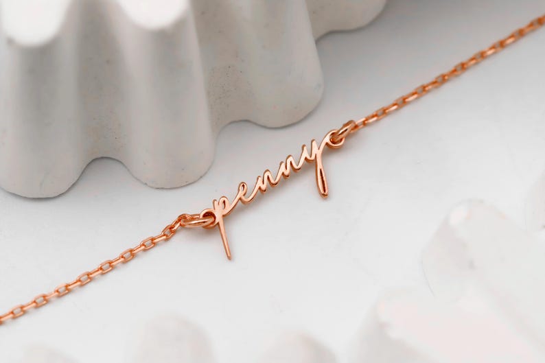 May include: A delicate rose gold necklace with the name "Penny" in cursive script. The chain is fine and links to the name pendant. The necklace is displayed on a white surface with a textured, wave-like background.