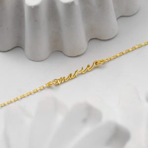May include: A delicate gold-colored necklace with the name "Marie" in cursive script. The necklace chain is a fine link design. The jewelry is displayed on a white surface with abstract decorative elements.