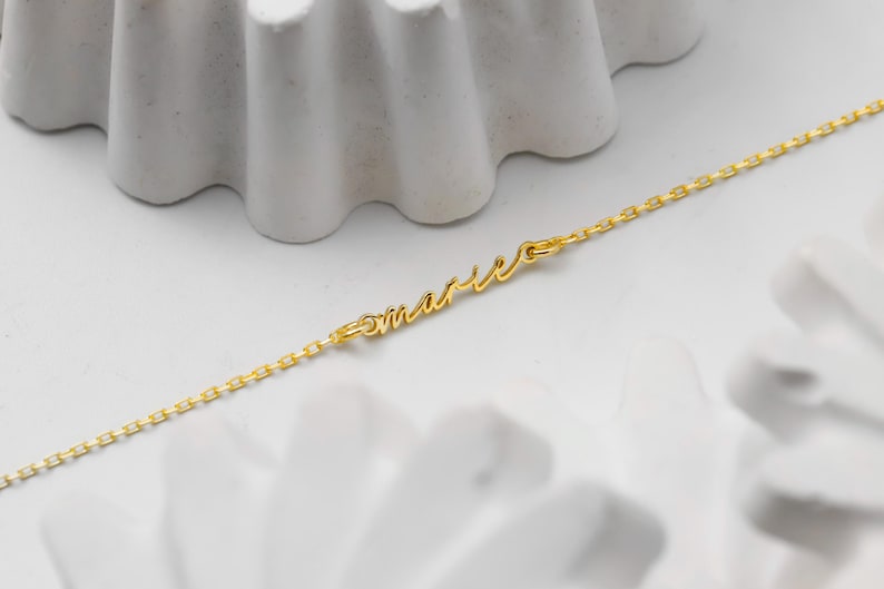 May include: A delicate gold-colored necklace with the name "Marie" in cursive script. The necklace chain is a fine link design. The jewelry is displayed on a white surface with abstract decorative elements.