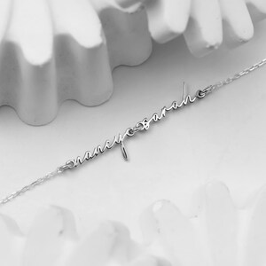 May include: A delicate silver necklace featuring the names "Nancy" and "Sarah" in elegant cursive script. The names are connected by a fine chain, creating a personalized and stylish accessory. The necklace is displayed against a soft, white background.