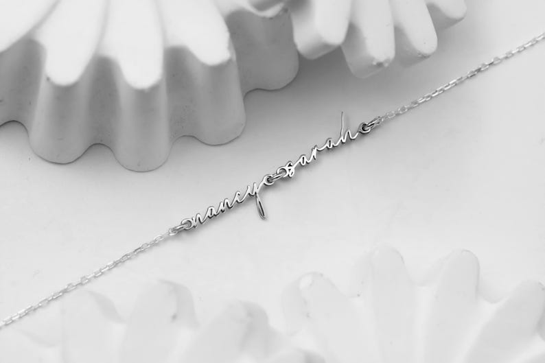 May include: A delicate silver necklace featuring the names "Nancy" and "Sarah" in elegant cursive script. The names are connected by a fine chain, creating a personalized and stylish accessory. The necklace is displayed against a soft, white background.