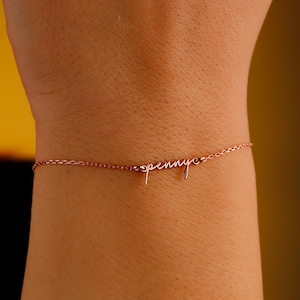 May include: Rose gold bracelet with the name "penny" in a delicate cursive script. The bracelet is a fine chain, and the name is the focal point of the jewelry. The bracelet is worn on a wrist.