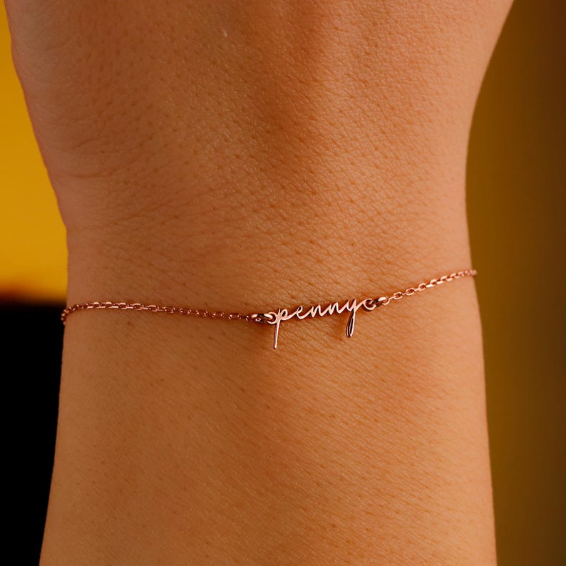 May include: Rose gold bracelet with the name "penny" in a delicate cursive script. The bracelet is a fine chain, and the name is the focal point of the jewelry. The bracelet is worn on a wrist.