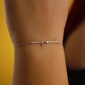 May include: A delicate silver bracelet with the names "nancy sarah" in cursive script. The bracelet is a fine chain with the names as a central design element. The background is a warm yellow.