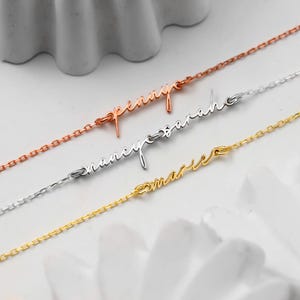 May include: Three delicate name necklaces in rose gold, silver, and gold. Each necklace features a cursive name: "Penny", "Nancy Sarah", and "Marie". The chains are fine and the names are elegantly scripted.