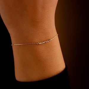 May include: A delicate gold-colored bracelet with the name "marie" in cursive script. The bracelet is a fine chain, and the name is spelled out in small, elegant letters. The bracelet is worn on a wrist.