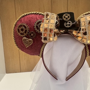 May include: Red and gold steampunk-themed Minnie Mouse ears with a black top hat and a white veil. The ears are decorated with gears, a heart, and a gold bow.