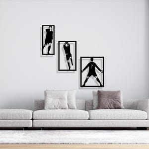 Può includere: Tre opere d'arte da parete in metallo nero raffiguranti calciatori in azione. Ogni figura è incorniciata in un bordo rettangolare. L'arte è montata su una parete bianca sopra un divano grigio.