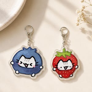 May include: Two clear acrylic keychains featuring cartoon cats. One cat is inside a blue berry shape, and the other is inside a red strawberry shape. Both charms have silver-toned metal lobster claw clasps attached to the top for hanging.