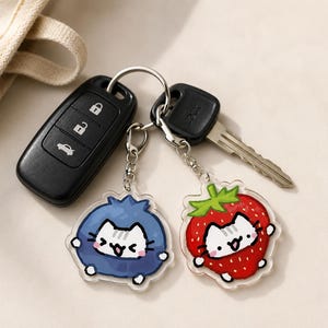 May include: Two acrylic keychains attached to a metal ring with a car key fob and a metal house key. One keychain features a white cat inside a blue blueberry, and the other features a white cat inside a red strawberry with green leaves.