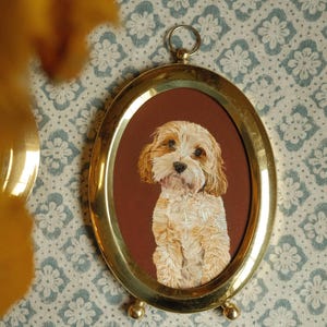 May include: An oval brass frame containing a detailed portrait of a light brown and white dog with floppy ears. The frame features a polished metallic finish, a small hanging loop at the top, and two small spherical feet at the base for standing.