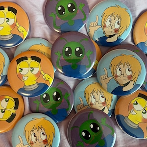 May include: A collection of circular pin-back badges featuring three distinct cartoon character designs: a yellow-skinned character with dark hair, a green-skinned alien with antennae, and a light-haired character wearing glasses and pointing upward.