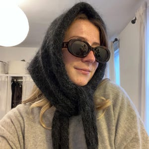 Può includere: Una persona indossa un maglione grigio e una sciarpa di lana nera. La sciarpa è avvolta intorno alla testa e al collo della persona. La persona indossa occhiali da sole ovali con lenti scure.