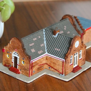 Puede incluir: Un edificio de ladrillo en miniatura con un tejado gris, un reloj y una puerta blanca. El edificio tiene adornos rojos y está sobre una base gris claro. Una pequeña planta en maceta blanca está en el fondo.