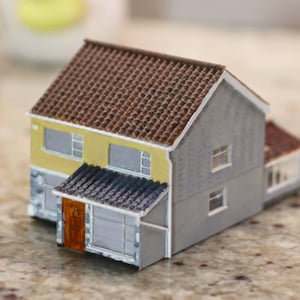 Könnte beinhalten: Miniatur-Hausmodell mit braunem Ziegeldach, gelben und grauen Wänden und einer kleinen Veranda. Das Haus hat mehrere Fenster und eine braune Tür. Das Modell steht auf einer gesprenkelten Oberfläche.