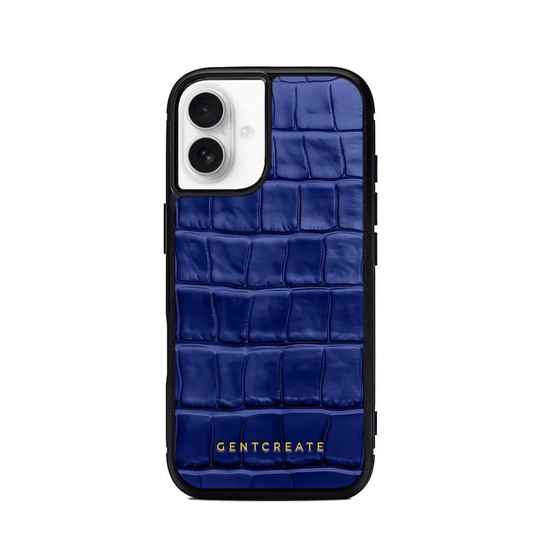 A crocodile-textured iPhone case. The case features a Glossy, reptile-inspired Glossy with raised scales that give it a luxurious, high-end look. The phone case has a black border, highlighting its sleek and modern design. The person's hand, with neatly manicured red nails, is seen against a backdrop of denim jeans, adding a stylish, fashionable touch to the image. The brand name "GENTCREATE" is subtly visible on the case.