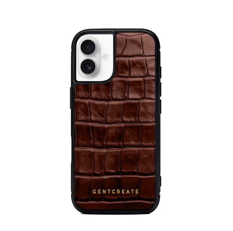 A crocodile-textured iPhone case. The case features a Glossy, reptile-inspired Glossy with raised scales that give it a luxurious, high-end look. The phone case has a black border, highlighting its sleek and modern design. The person's hand, with neatly manicured red nails, is seen against a backdrop of denim jeans, adding a stylish, fashionable touch to the image. The brand name "GENTCREATE" is subtly visible on the case.