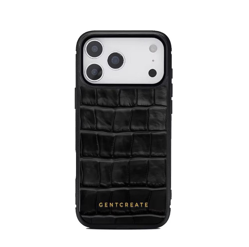 A crocodile-textured iPhone case. The case features a Glossy, reptile-inspired Glossy with raised scales that give it a luxurious, high-end look. The phone case has a black border, highlighting its sleek and modern design. The person's hand, with neatly manicured red nails, is seen against a backdrop of denim jeans, adding a stylish, fashionable touch to the image. The brand name "GENTCREATE" is subtly visible on the case.